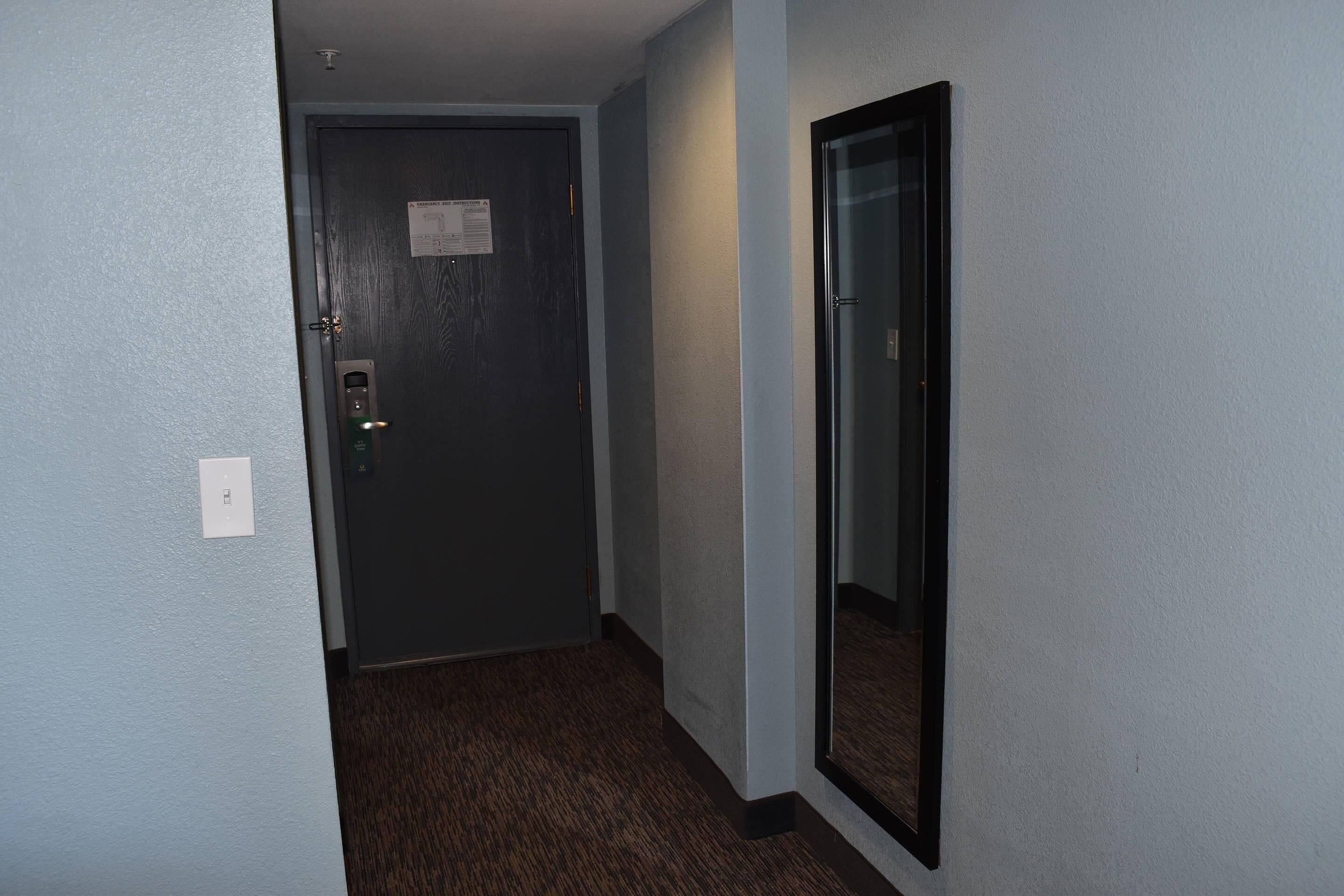 A narrow hallway with a door at the end, a light switch on the wall, and a mirror on the right.