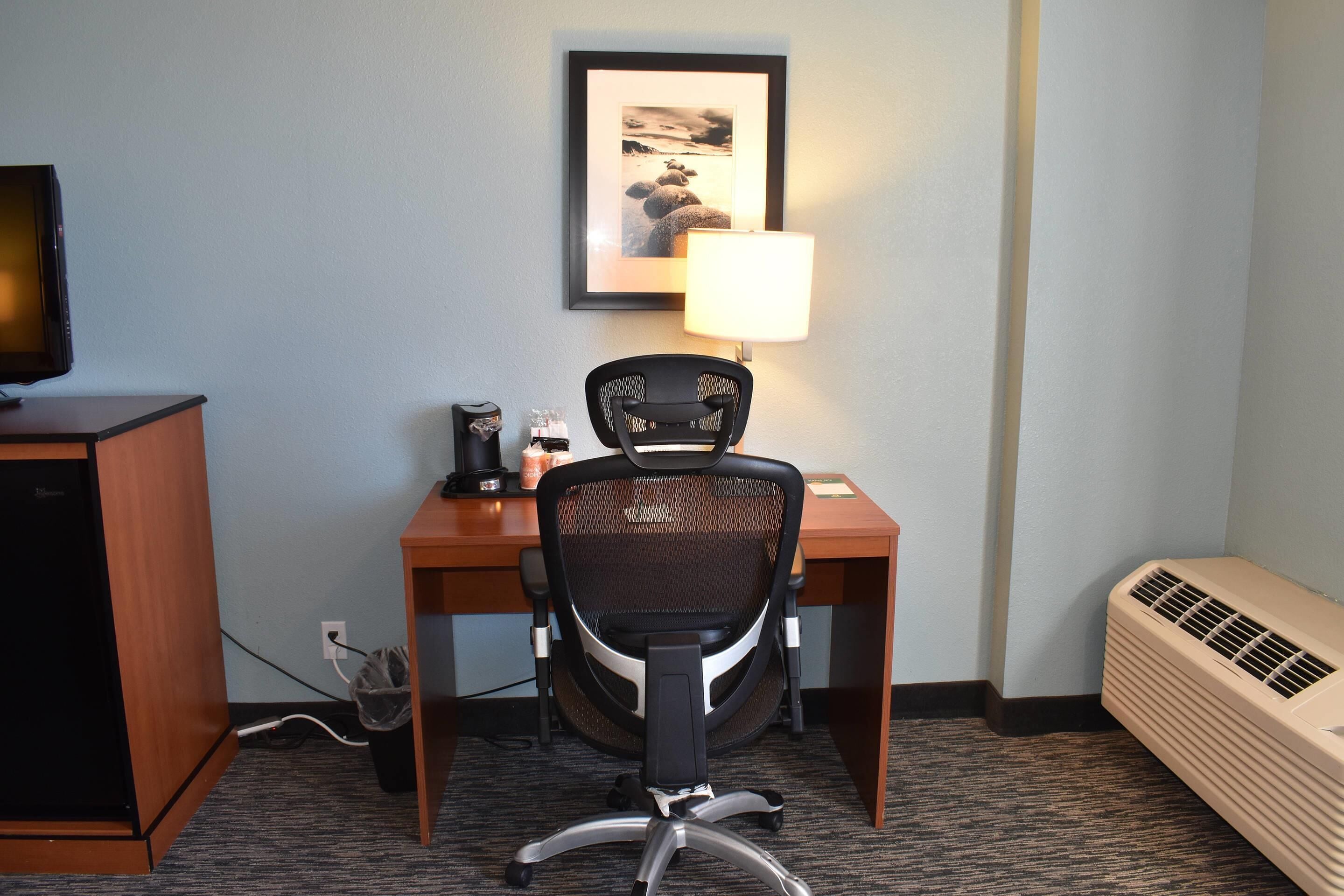 A hotel room with a desk, chair, lamp, air conditioner, and a television on a cabinet.