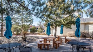 An outdoor patio area with round tables and benches, surrounded by trees and a house in the background. - Quality Inn and Suites Garden of the Gods (Colorado Springs)