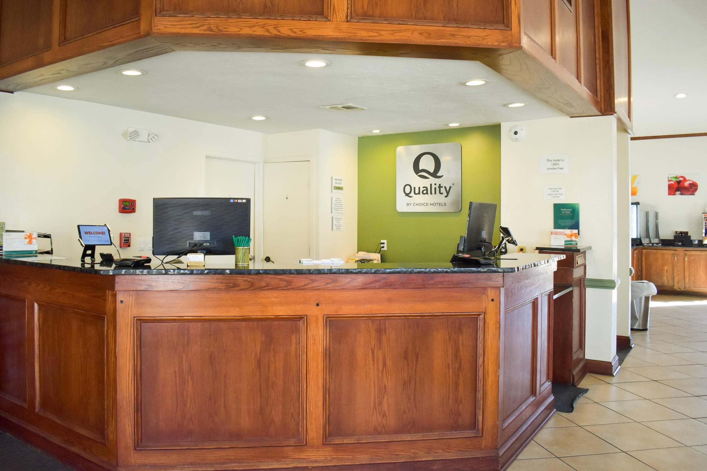 A hotel reception area with a wooden reception desk, a green wall with the hotel's logo, and a computer monitor.