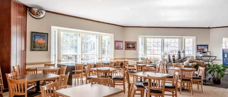 A spacious dining area with multiple wooden tables and chairs, large windows allowing natural light, and framed artwork on the walls.