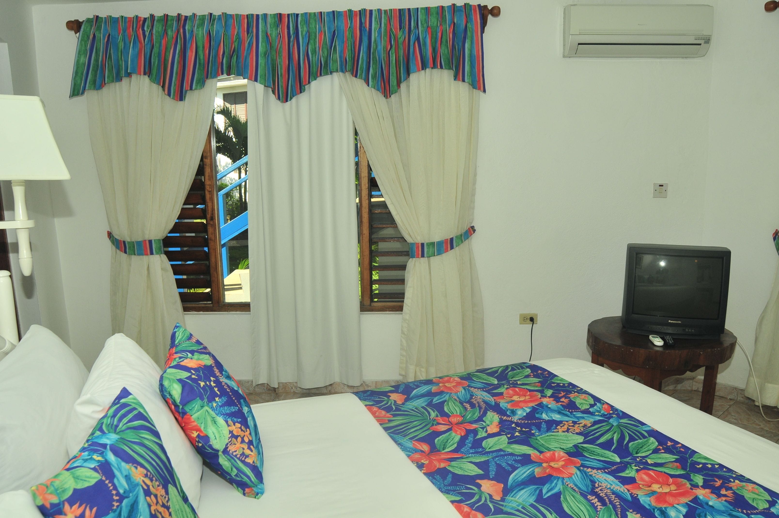 Quarto com cama, almofadas, cortinas, uma televisão e um ar condicionado.