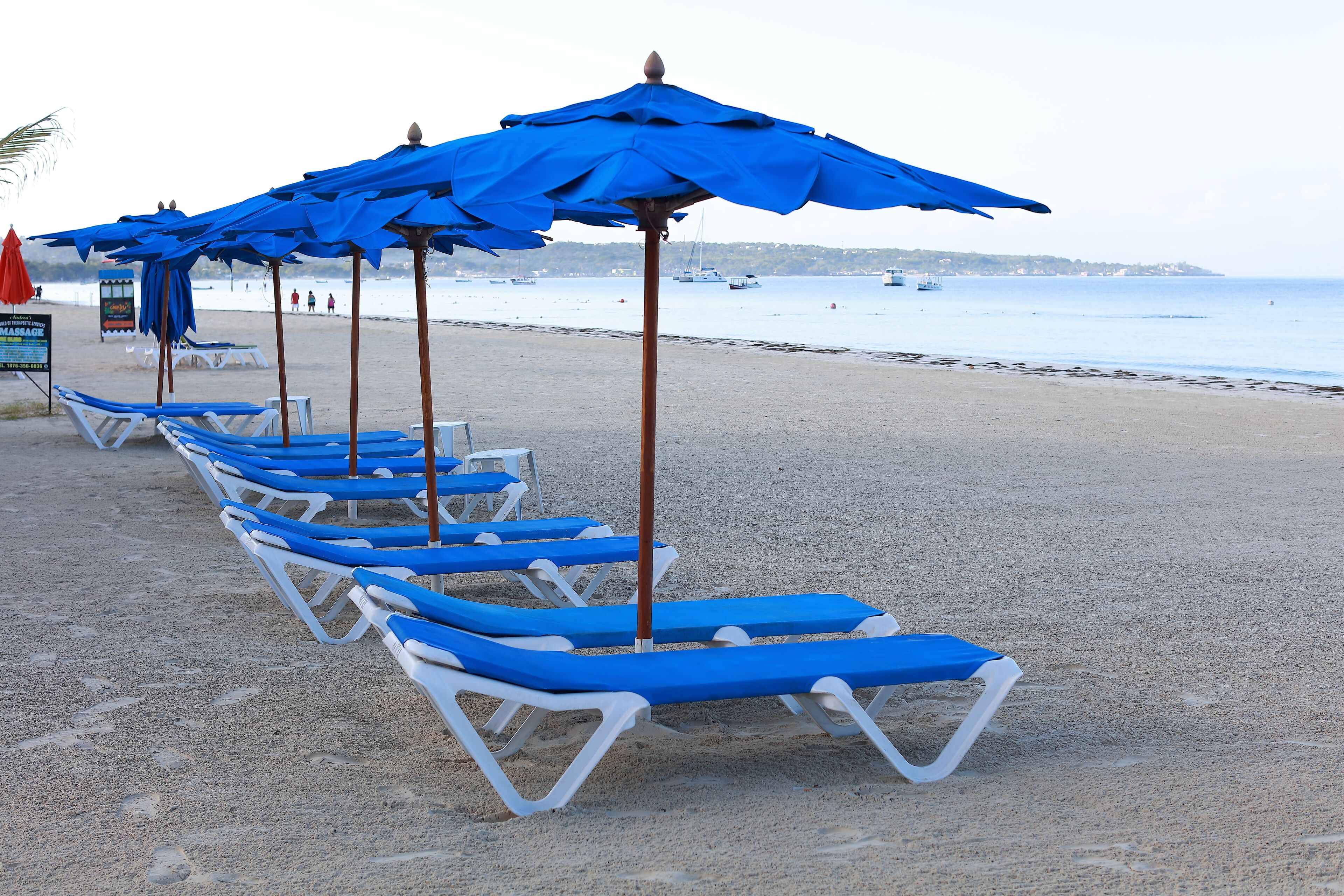A beach with rows of blue sun loungers and matching umbrellas, a signboard, and boats in the background.