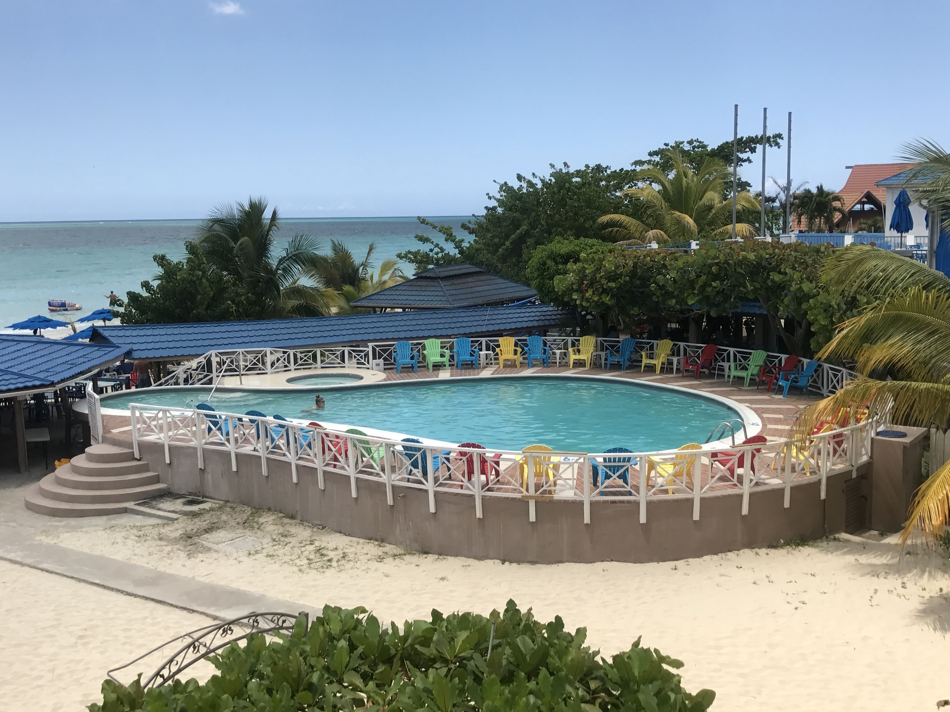 Uma área de piscina com uma piscina circular, rodeada por cadeiras e guarda-sóis coloridos, e vista para o oceano e a praia ao fundo.