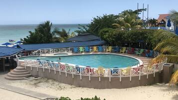Uma área de piscina com uma piscina circular, rodeada por cadeiras e guarda-sóis coloridos, e vista para o oceano e a praia ao fundo.