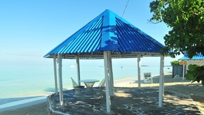 A beach gazebo with a blue roof, a table, and chairs, surrounded by a sandy area with a sign indicating a house hotel.