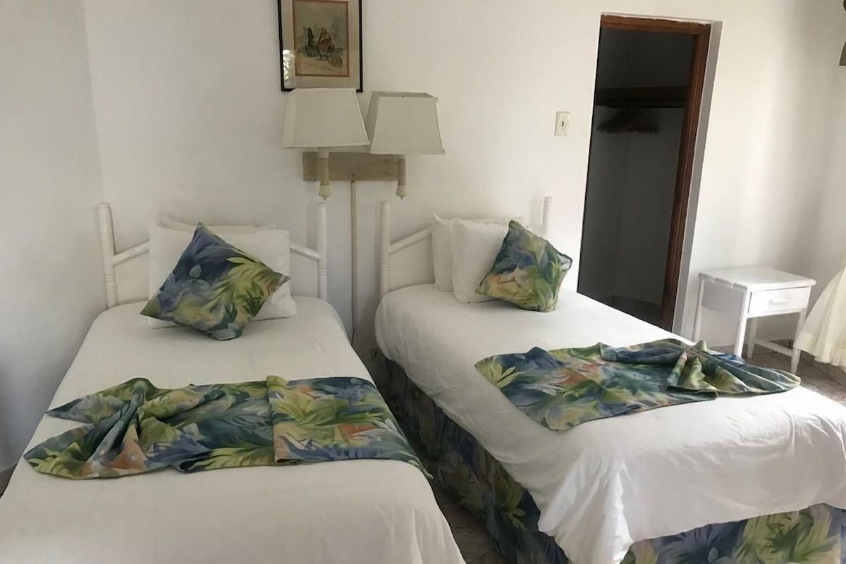 Quarto com duas camas, cada uma com colcha florida e almofadas combinando. Pequeno criado-mudo com abajur em cada cama.