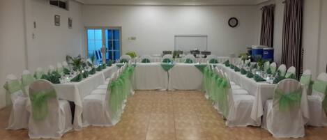 Un salón de conferencias con mesas dispuestas para una reunión, sillas con cintas verdes, un reloj en la pared y una ventana con persianas.