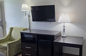 A room with a flat-screen TV mounted on the wall, a desk with drawers, a green patterned chair, and two table lamps.
