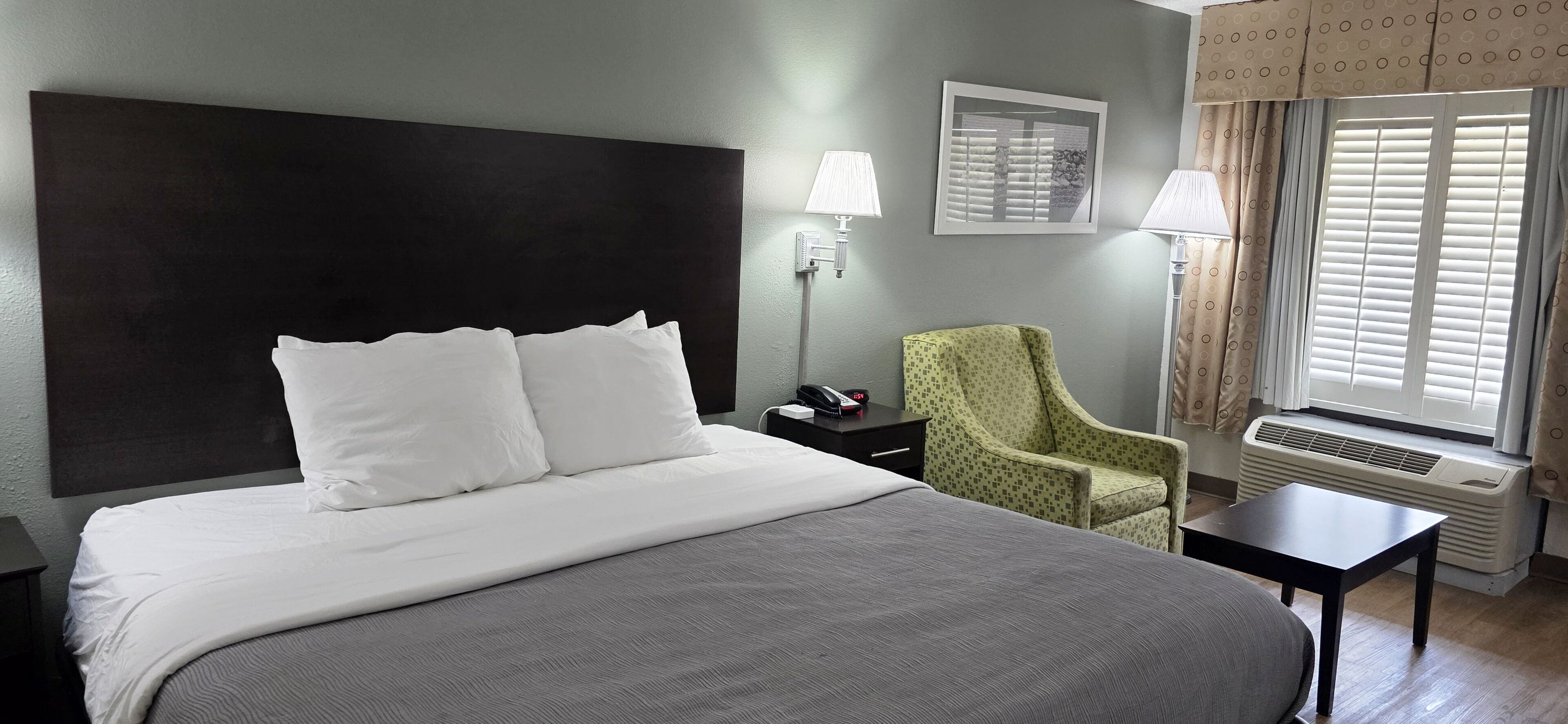 A hotel room with a large bed, a chair, a bedside table, a lamp, a picture on the wall, and a window with curtains.