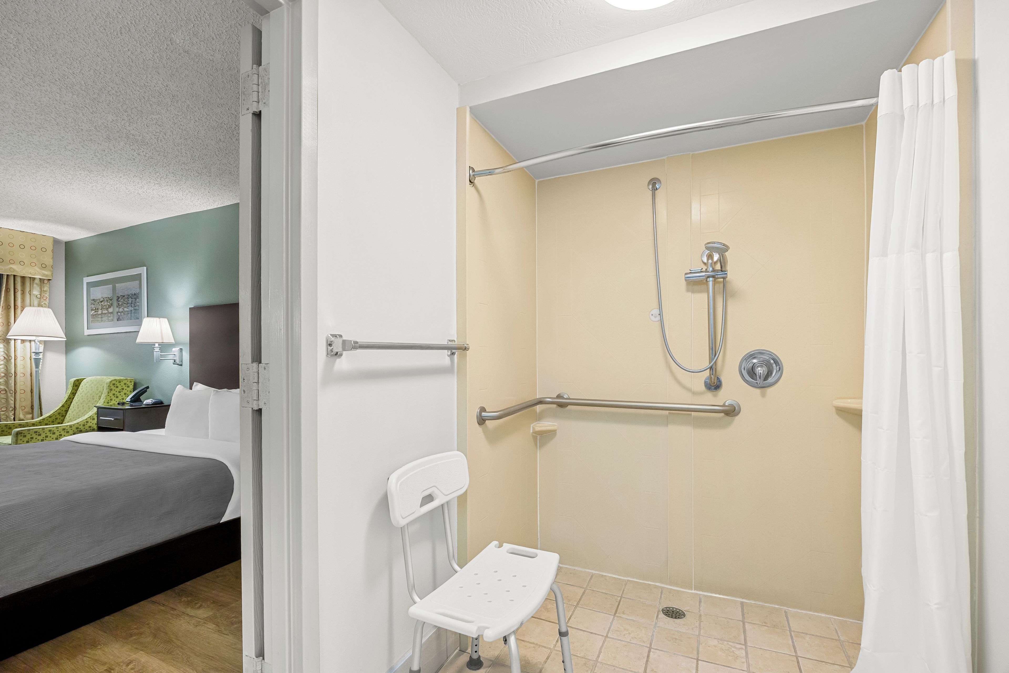 Une salle de bain d’hôtel équipée d’une douche, d’une barre d’appui, d’une poignée de maintien et d’une petite chaise blanche.