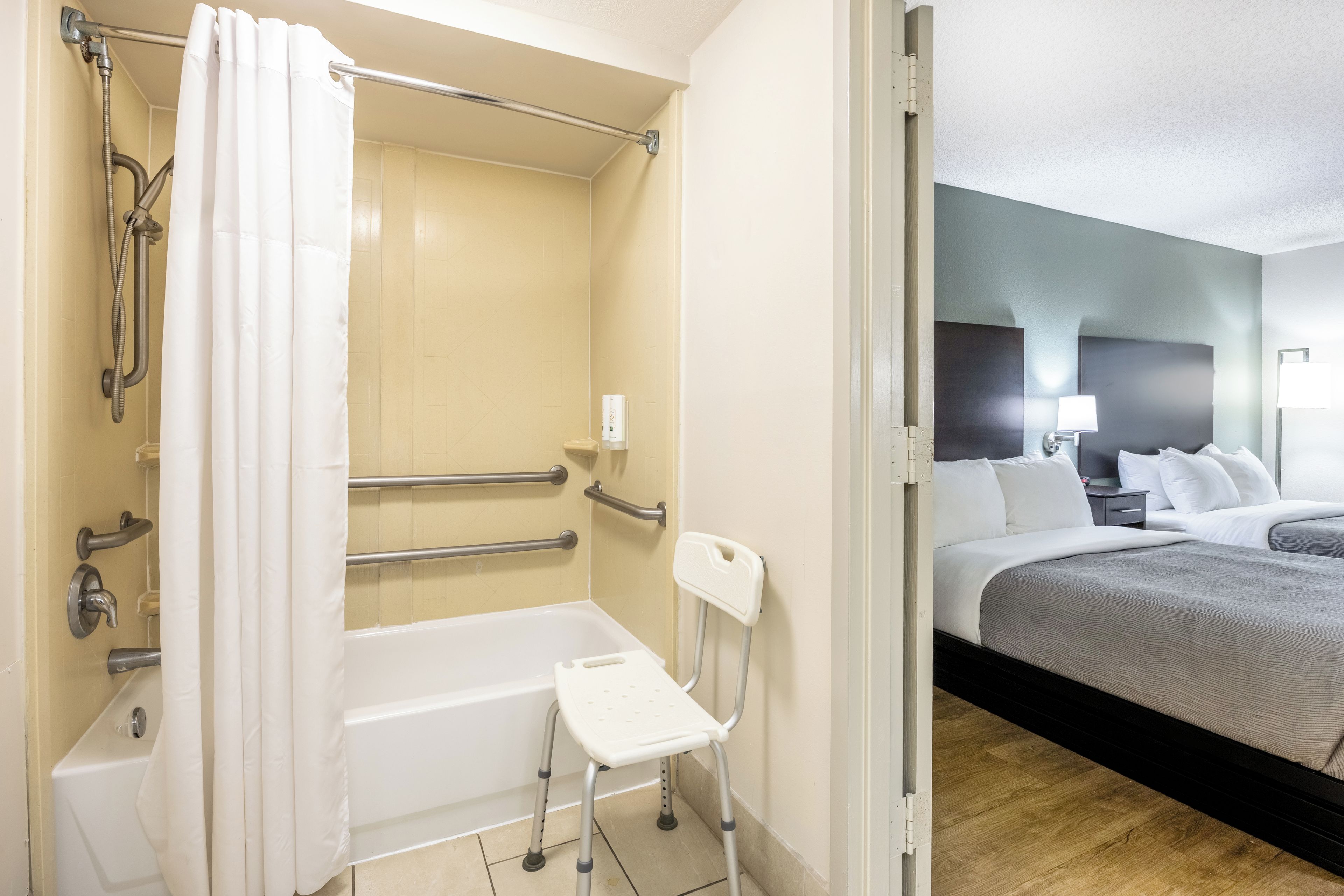 A hotel bathroom with a walk-in shower, grab bars, a white toilet, and a small white chair.