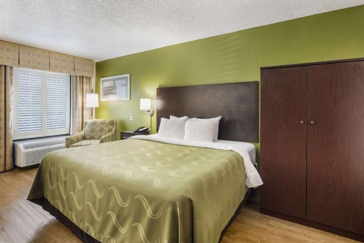 A hotel room with a bed, a wardrobe, a chair, a desk with a lamp, and a window with curtains.