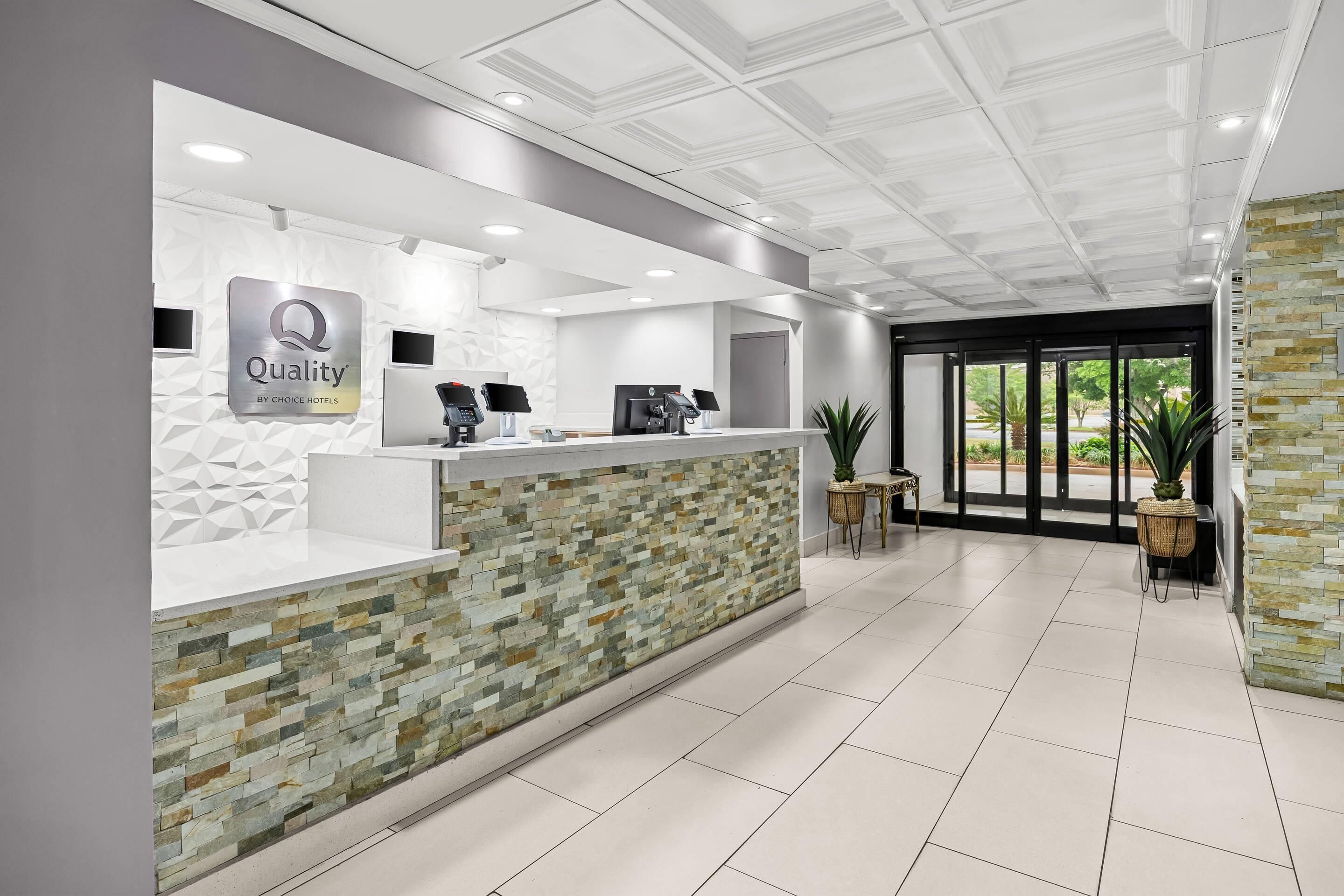 Zona de recepción de un hotel con diseño moderno, que incluye un mostrador de recepción, una pantalla digital y un cartel que dice 'Quality by Choice Hotels'.