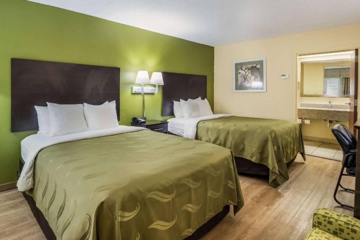 A hotel room with two beds, a desk, a chair, a mirror, and a wall-mounted lamp.