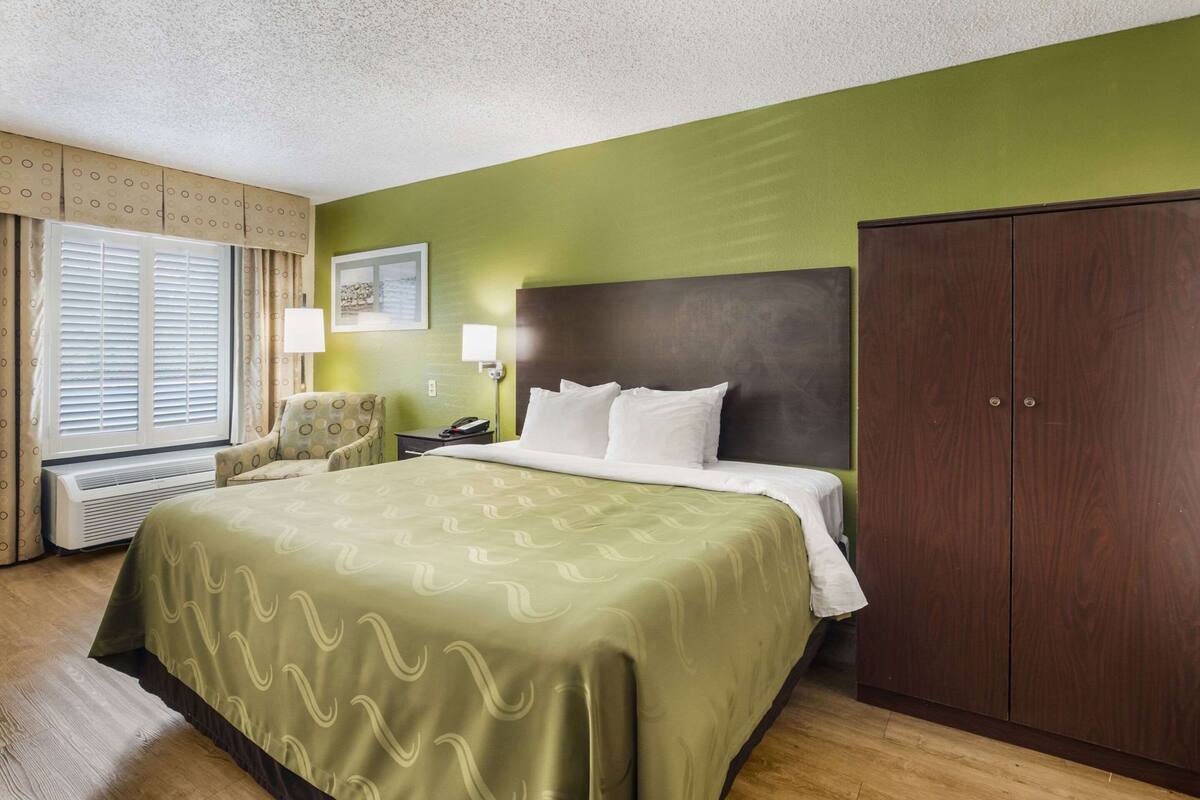 A hotel room with a bed, a chair, a wardrobe, and a window with curtains.