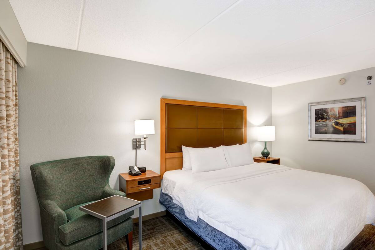 Una habitación de hotel con una cama, un sillón verde, una mesita, dos lámparas de noche y un cuadro en la pared.