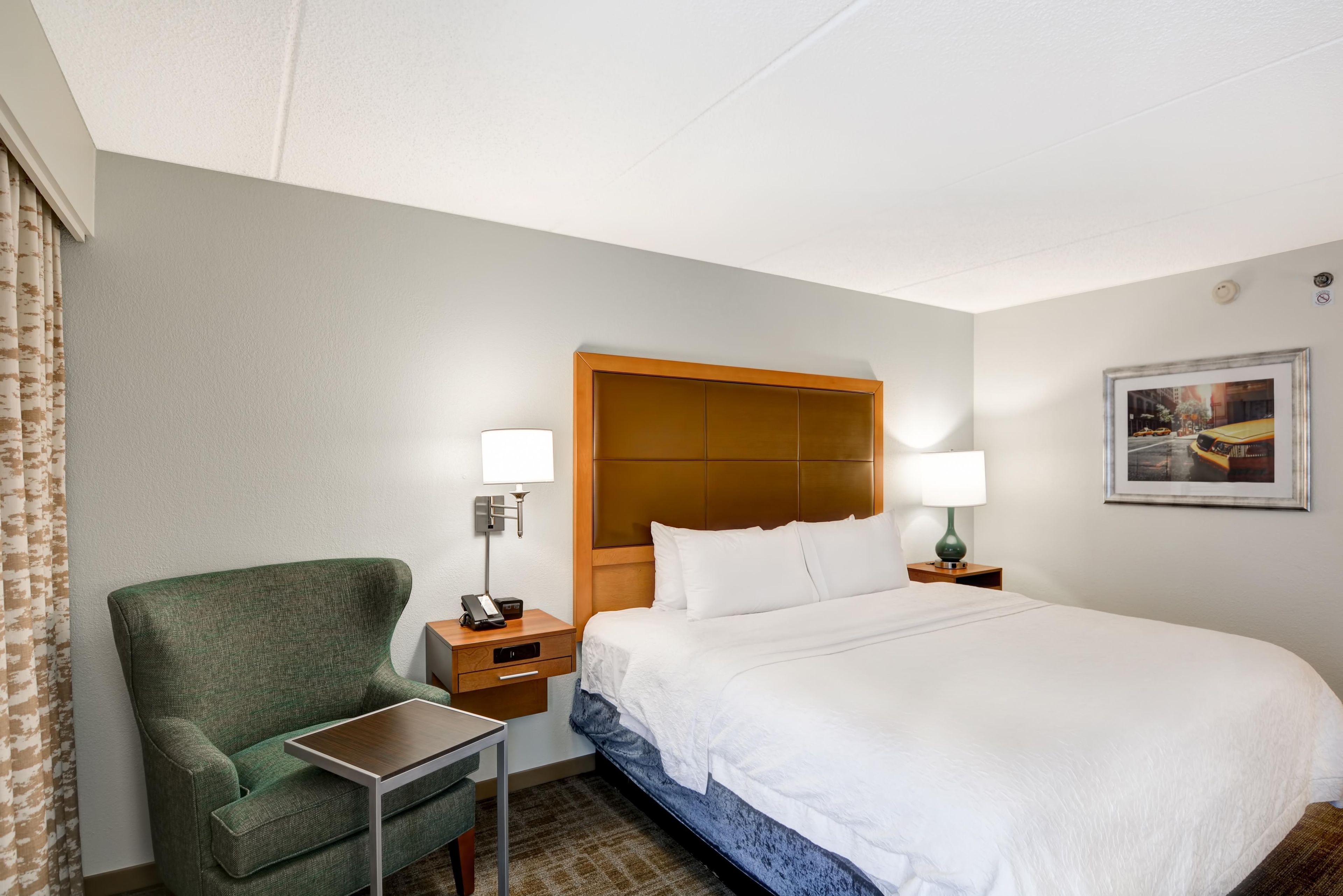 A hotel room with a bed, a green armchair, a small table, a nightstand with a lamp, and a framed picture on the wall.