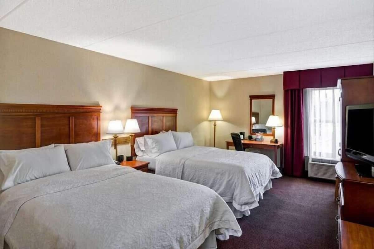 Una habitación de hotel con dos camas, un escritorio con silla, un televisor y una ventana con cortinas.