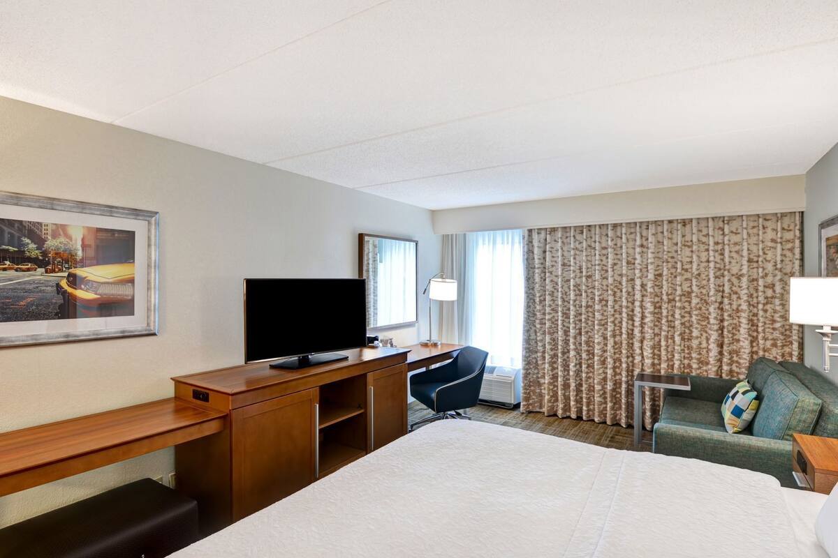 Habitación de hotel con cama, escritorio con televisor, silla, mesita pequeña y ventana con cortinas.