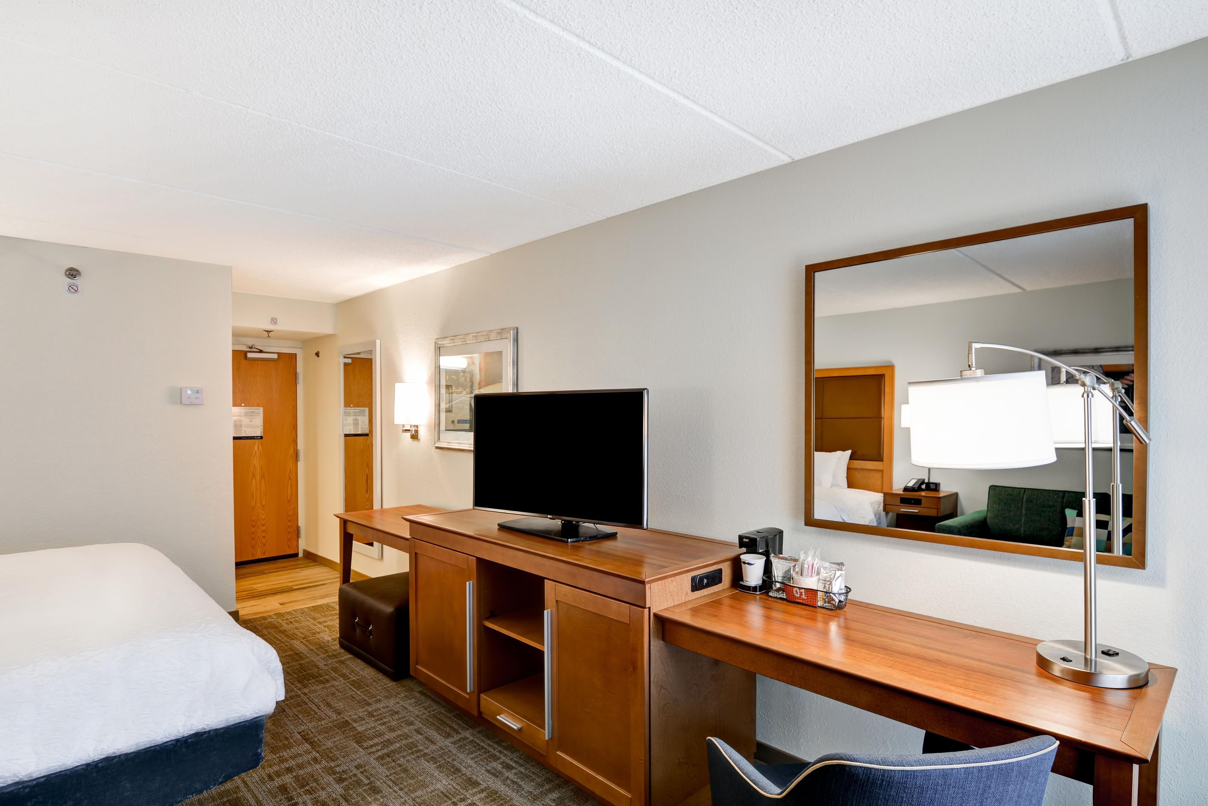 A hotel room with a bed, a desk with a television, a mirror, a coffee maker, and a chair.