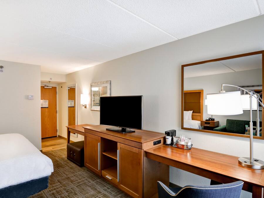 A hotel room with a bed, a desk with a television, a mirror, a coffee maker, and a chair.