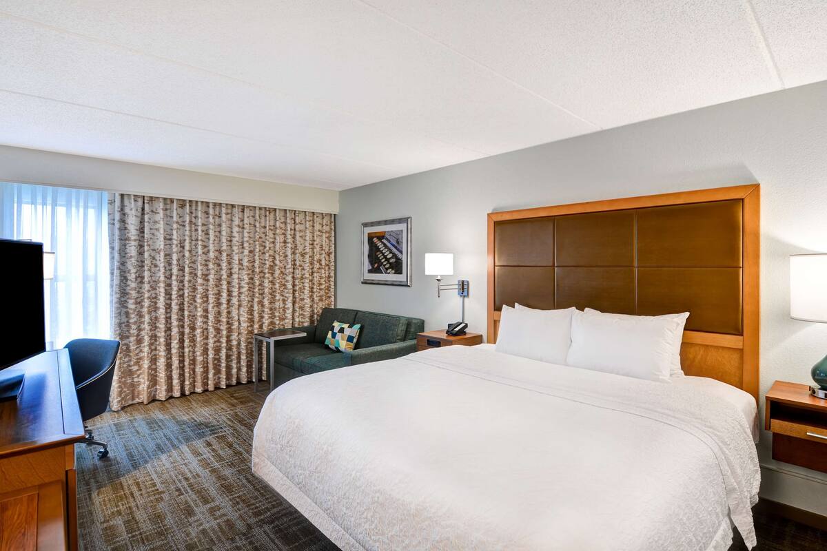 Habitación de hotel con una cama grande, un escritorio con computadora, una silla, un sofá y una ventana con cortinas.