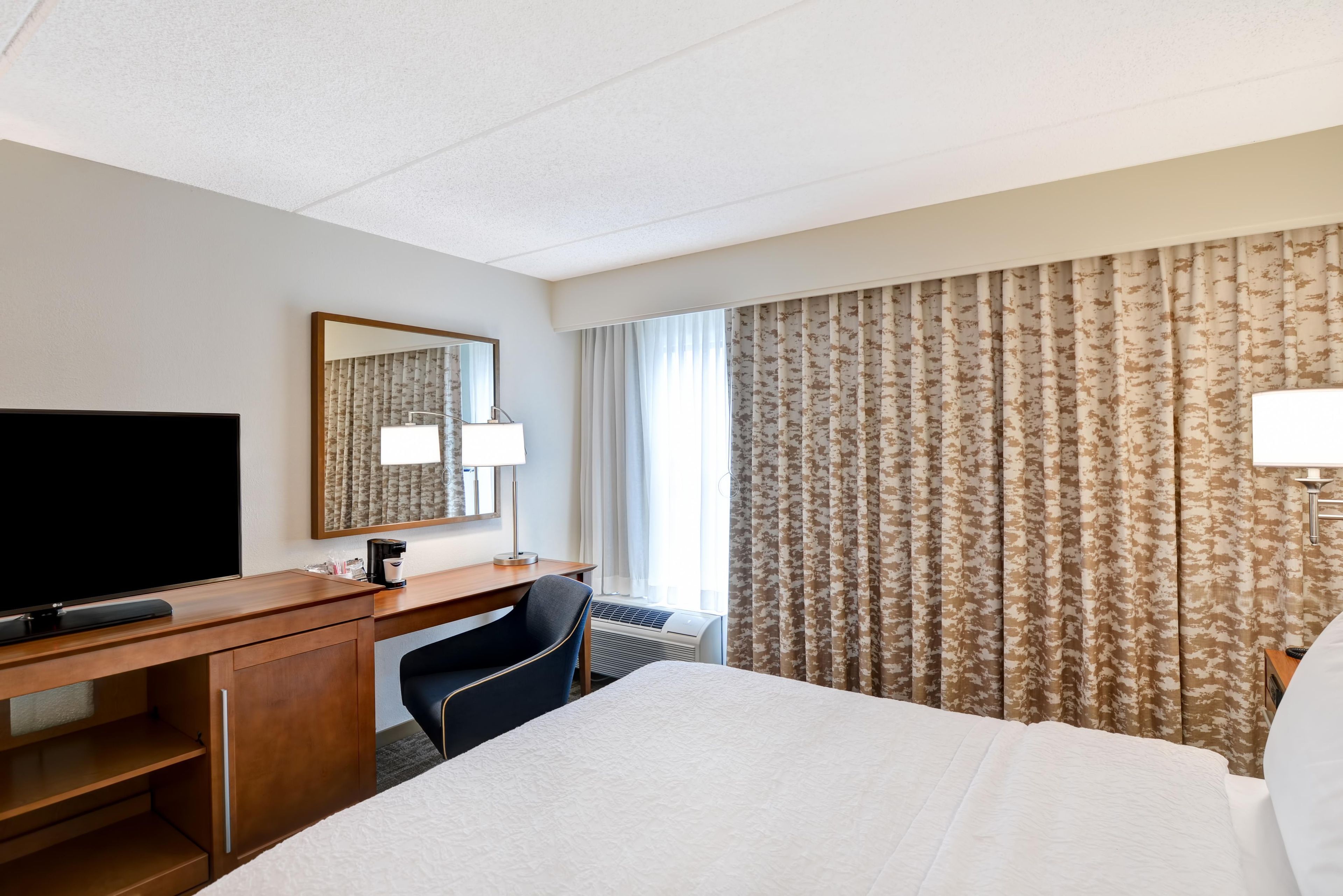Une chambre d’hôtel comprenant un lit, un bureau avec une télévision, une chaise, un miroir et des rideaux.