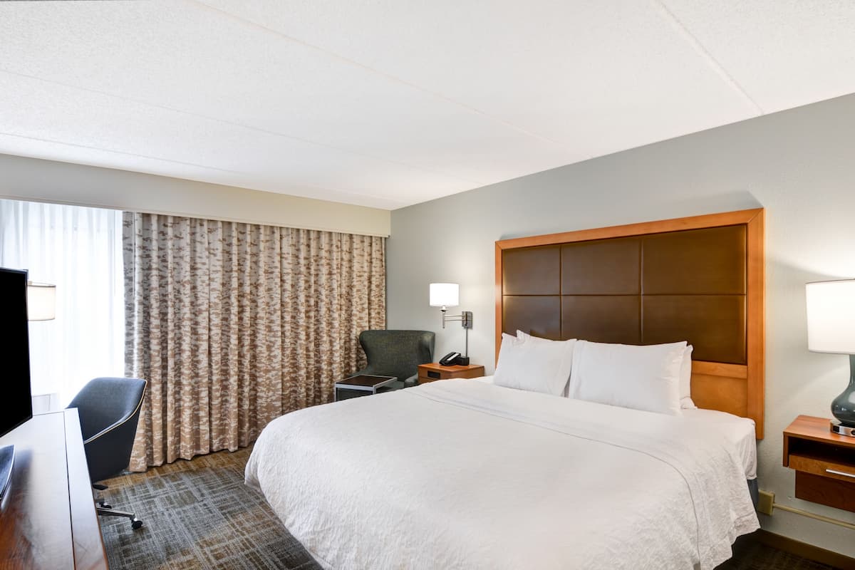 Habitación de hotel con una cama grande, un escritorio con silla, una mesita de noche con lámpara y una ventana con cortinas.