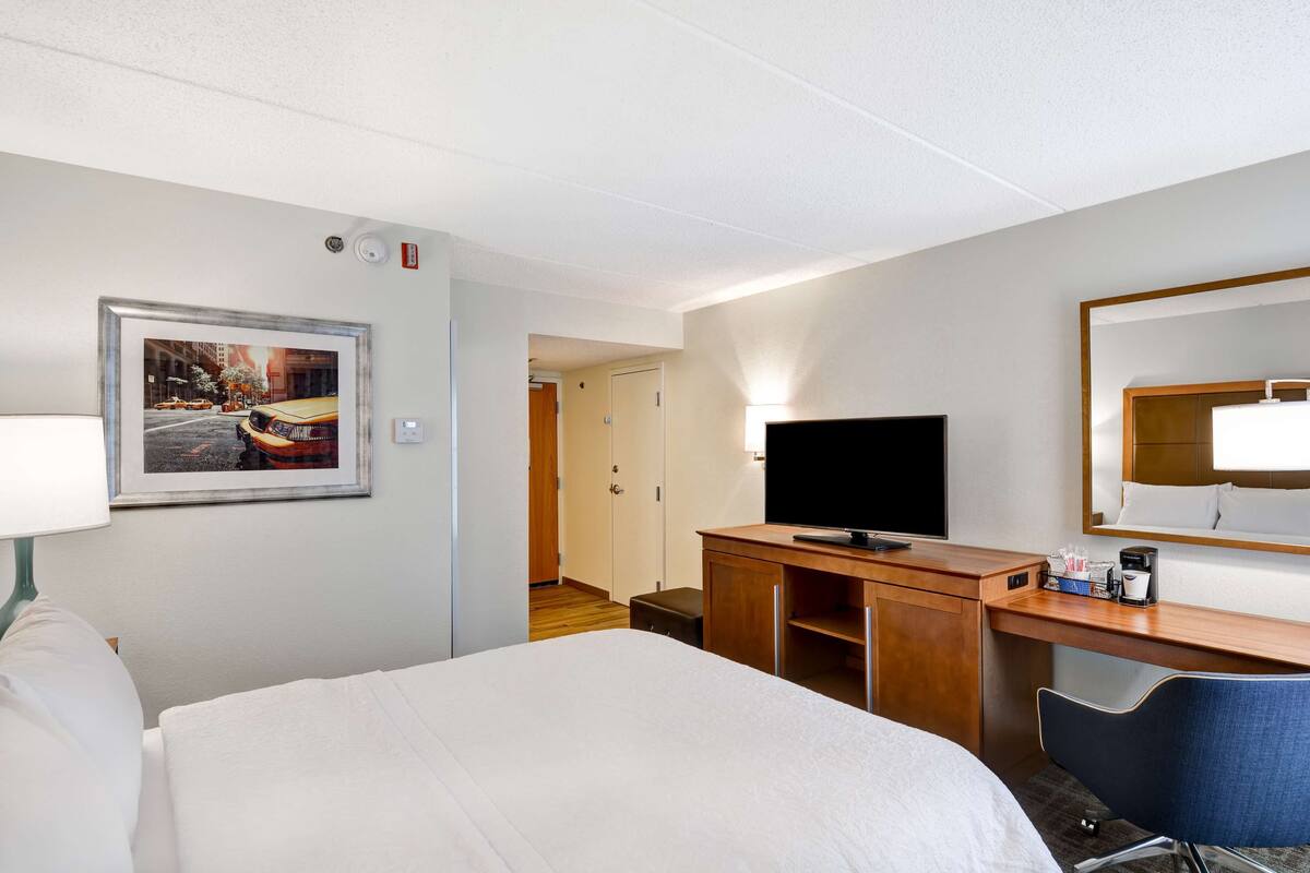 Un cuarto de hotel con una cama, un escritorio con televisor, una silla, un cuadro en la pared y una puerta que da a otra habitación.