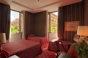 Una camera d'hotel con due letti, una scrivania, una sedia, una lampada e vista su edifici e alberi.