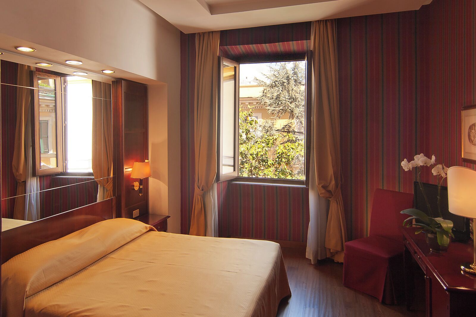A hotel room with a bed, a desk with a lamp, a chair, a window with curtains, and a view of trees outside.