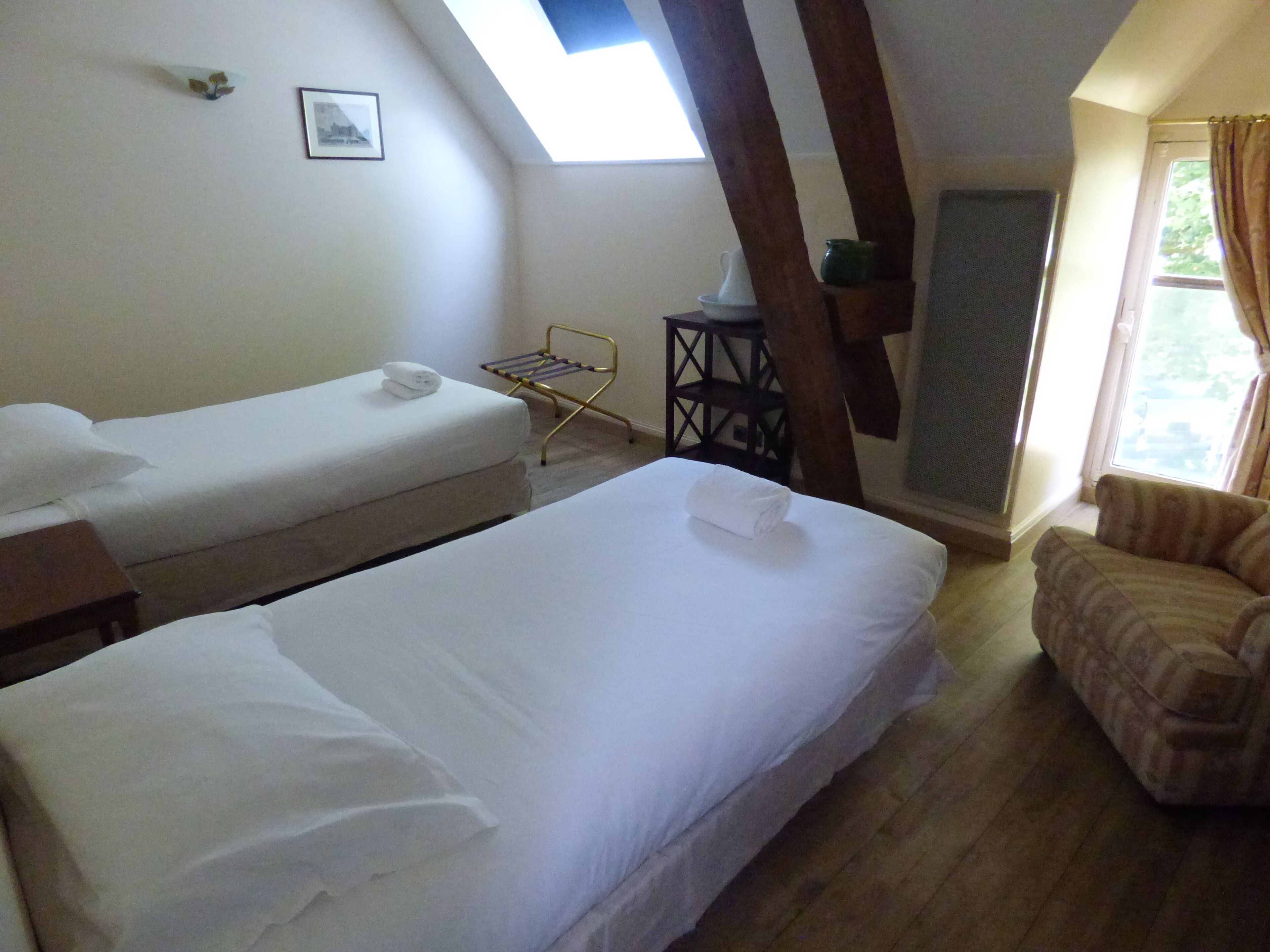A room with two single beds, a small table, a chair, a window, and a picture on the wall.