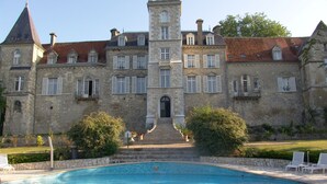 Un gran edificio de piedra con una torre central, una piscina delantera y un jardín bien cuidado.