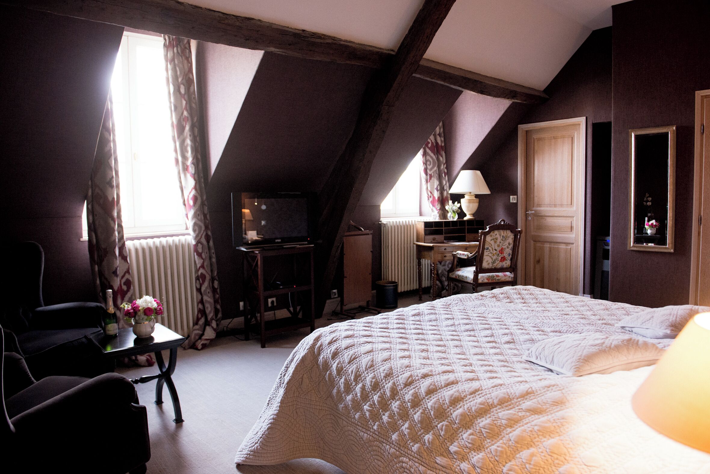 A bedroom with a large bed, a desk with a chair, a radiator, and a window with curtains.