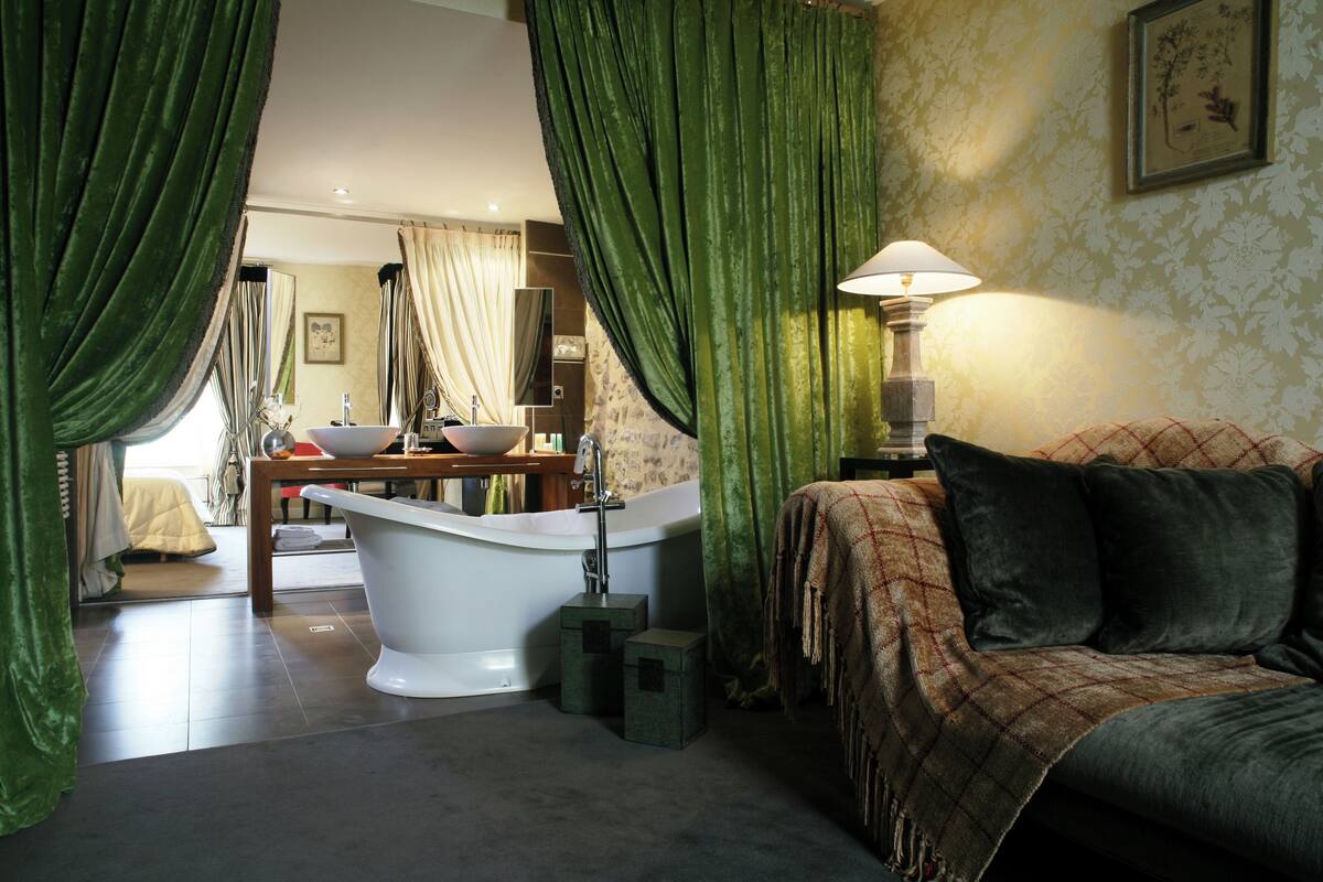 Een badkamer met een bad, een wastafel en een spiegel. Er is een groen gordijn en een lamp op een nachtkastje.