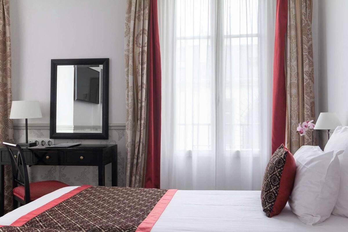 A hotel room with a bed, a desk, a chair, a mirror, and curtains.