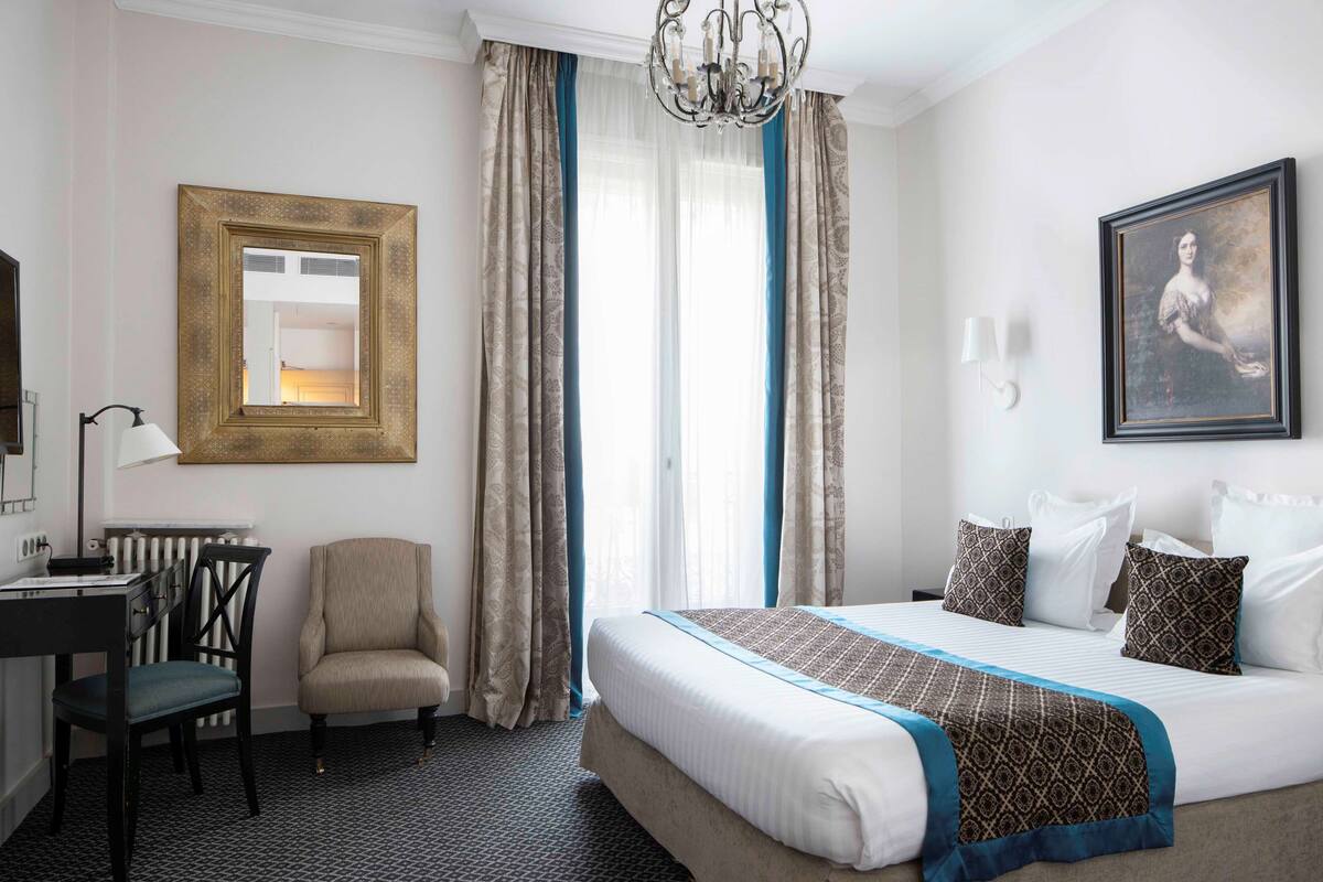 A hotel room with a bed, a desk, a chair, a radiator, and a chandelier.