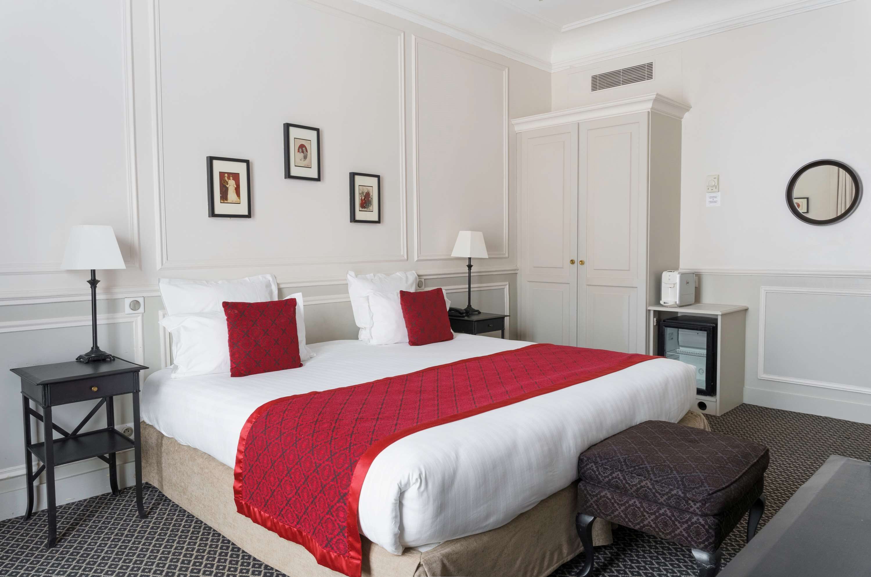 Une chambre d’hôtel avec un grand lit, deux tables de chevet avec des lampes, un petit radiateur, un miroir et des tableaux encadrés accrochés au mur.