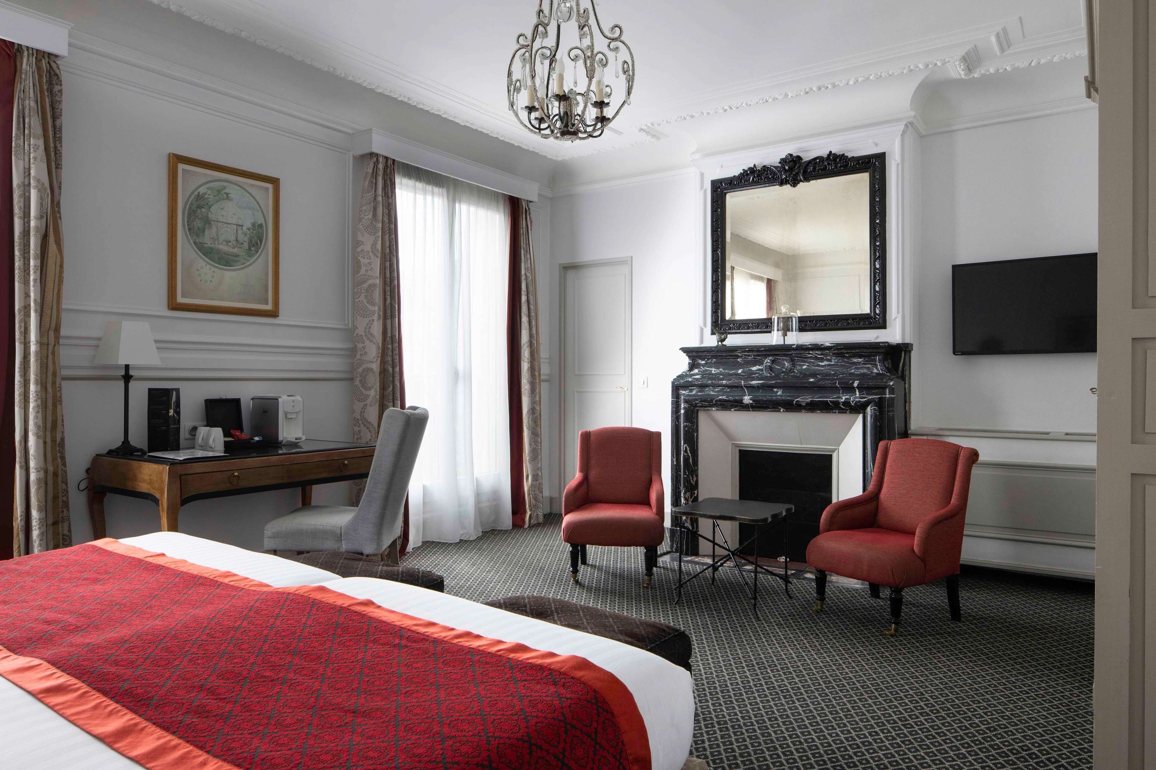 A hotel room with a large bed, two armchairs, a desk with a computer, a fireplace, and a TV on the wall.