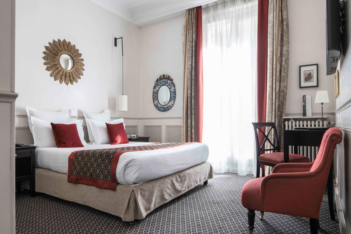 A hotel room with a large bed, a red armchair, a mirror, and a radiator.