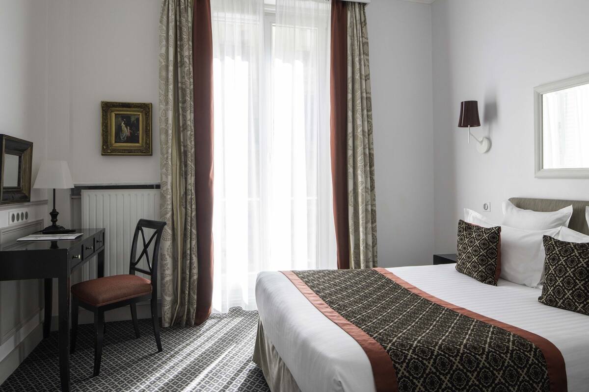 A hotel room with a bed, a desk, a chair, a lamp, and a window with curtains.