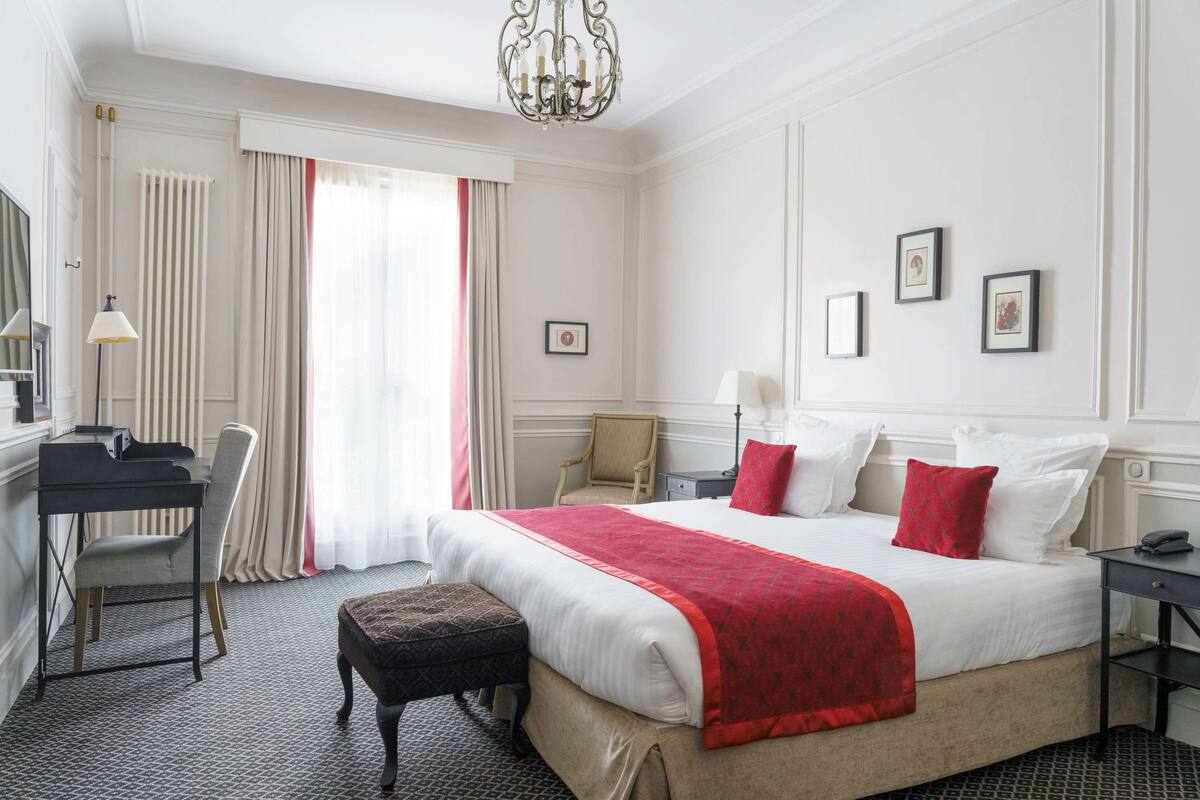 A hotel room with a large bed, a desk, a chair, a small table, and a bench.