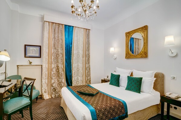 Una habitación de hotel con una cama grande, un escritorio con silla, una araña, un espejo y una ventana con cortinas.