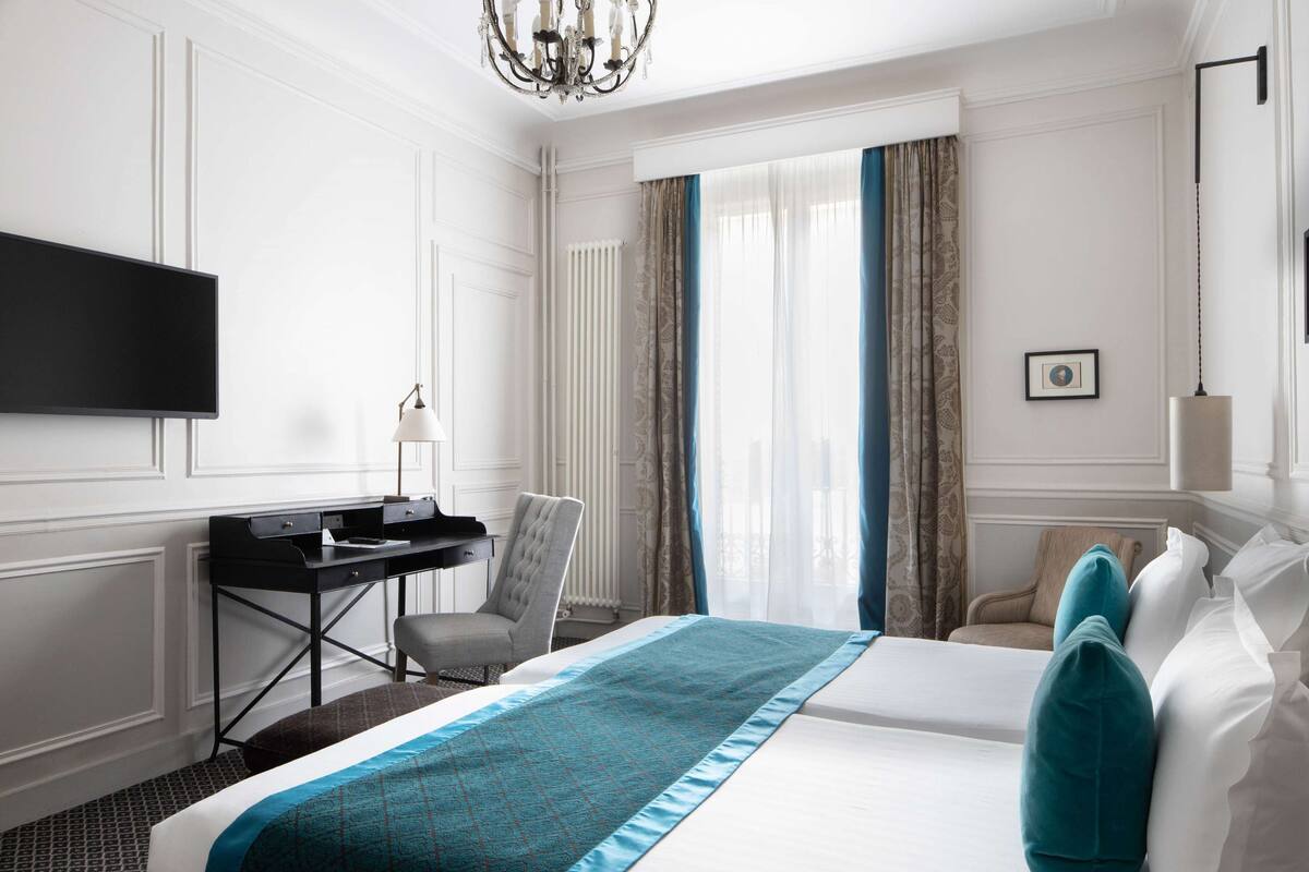 A hotel room with a bed, a desk, a chair, a TV, and a chandelier.