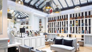 A spacious living area with a high ceiling, a central seating area with a sofa and chairs, a kitchen counter with various items, and a bookshelf filled with books and decorative objects. - Hôtel Bradford Elysées – Astotel (Paris)