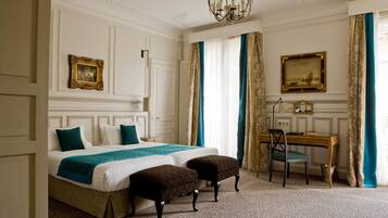 A hotel room with a bed, two ottomans, a desk, a chair, a chandelier, and two paintings on the wall.