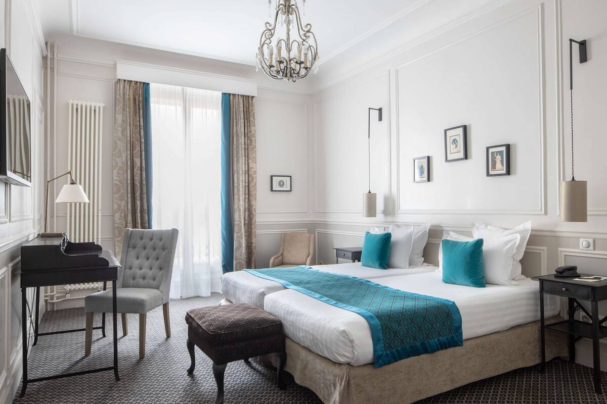 A hotel room with a piano, a tufted armchair, a bed with blue bedding, and a chandelier.