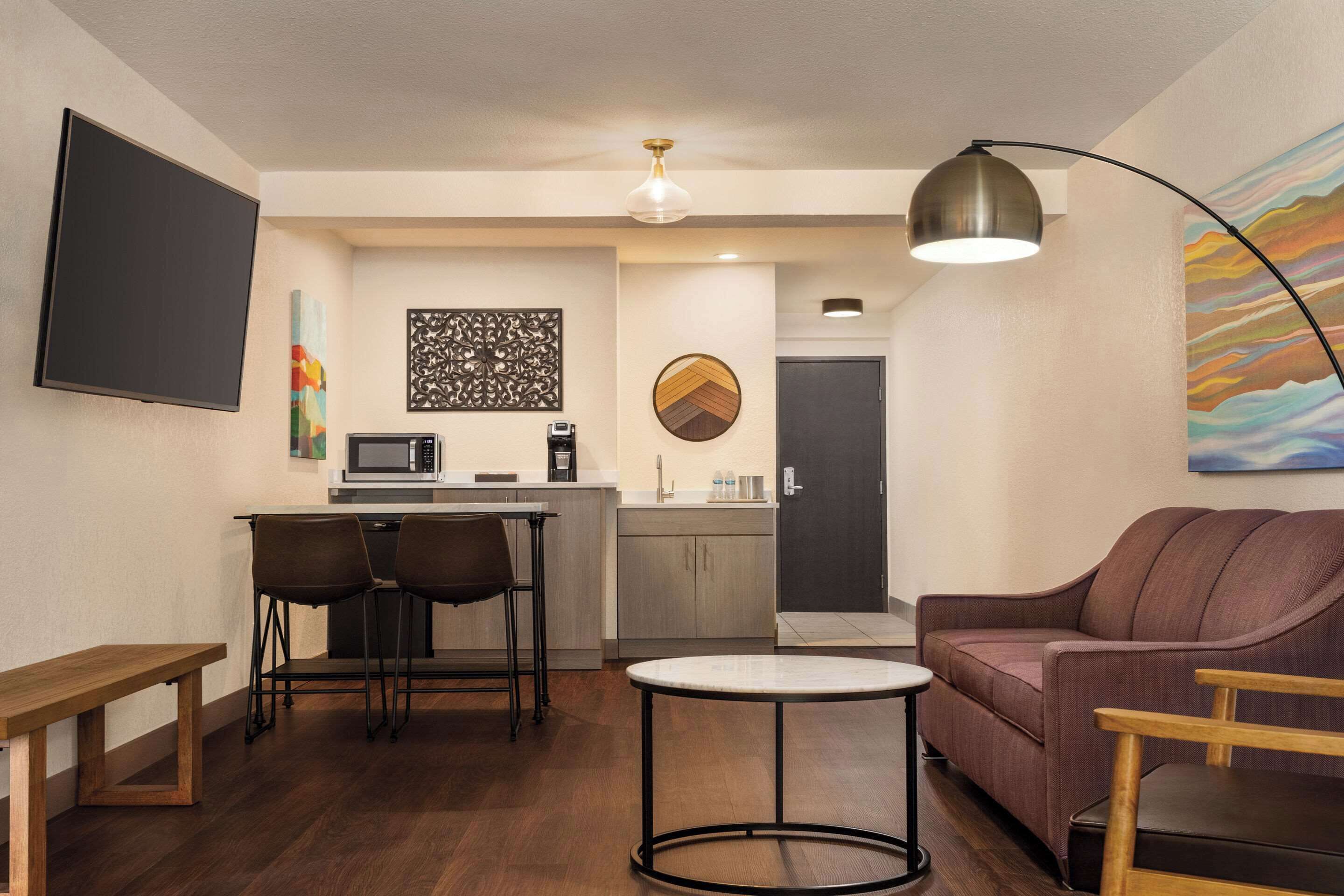 A modern living room with a sofa, a round coffee table, a TV, a kitchen area with a microwave, and a dining table with chairs.
