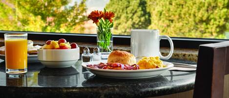 A breakfast table with a glass of orange juice, a bowl of fruit, a plate of eggs, toast, and a mug, set against a scenic view of trees and mountains.